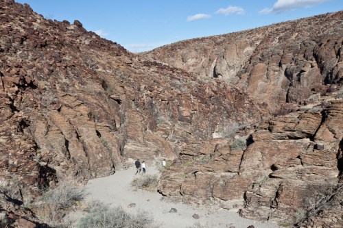 Two hikers navigate a rocky canyon landscape with dry vegetation under a clear blue sky.