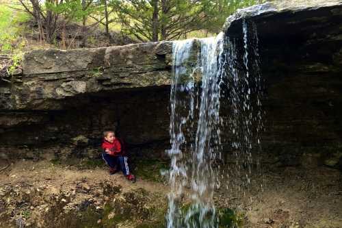 A young boy sits beside a small waterfall, surrounded by rocky terrain and greenery.