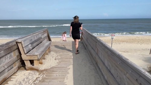 A woman walks down a sandy path towards the beach, while a child plays nearby. The ocean is visible in the background.