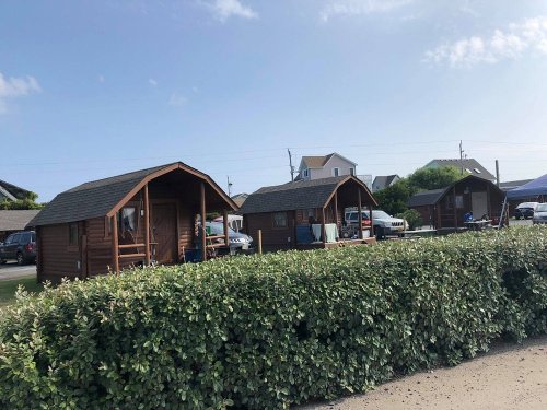 Three wooden cabins lined up near a hedge, with cars parked nearby and a clear blue sky above.