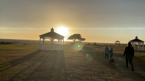 A sunset over the ocean with silhouettes of people walking a dog near gazebos on a grassy shore.