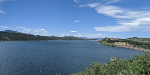 A panoramic view of a serene lake surrounded by mountains and blue skies with scattered clouds.