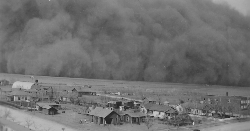 A historic black-and-white photo of a dust storm approaching a small town, with houses and trees in the foreground.