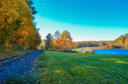 A scenic view of a railway track beside a lake, surrounded by colorful autumn trees and a clear blue sky.