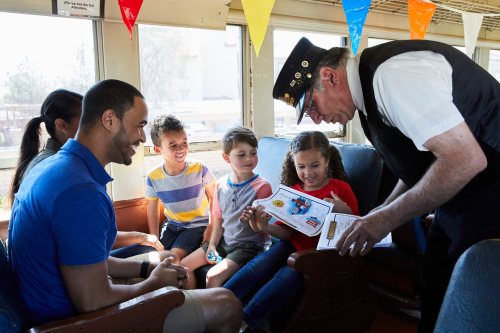 A train conductor shows a group of children a booklet while a man smiles at them in a colorful train interior.