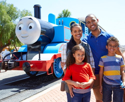 A smiling family poses in front of a large blue train character at a theme park.