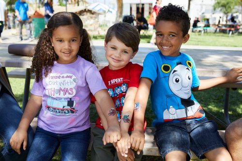 Three children sit together on a bench, smiling and holding hands, wearing colorful t-shirts in a sunny park setting.