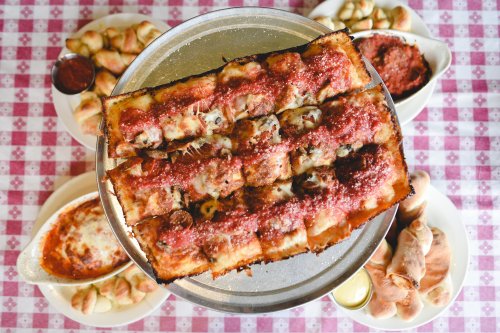 A large tray of cheesy, saucy pizza with garlic knots and baked pasta dishes on a checkered tablecloth.