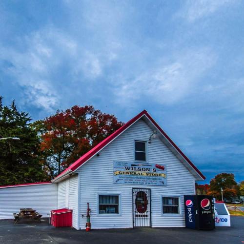 A white general store with a red roof, surrounded by trees and a cloudy sky, featuring a Pepsi vending machine outside.