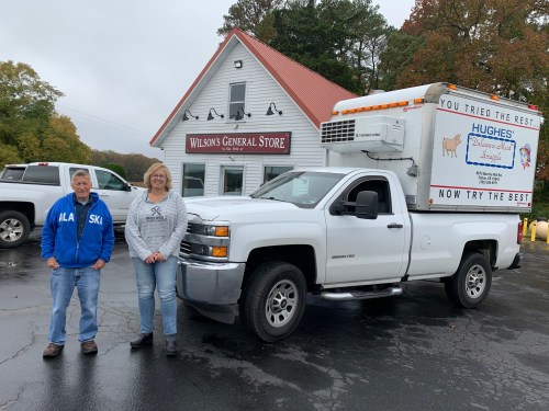 Two people stand beside a white pickup truck in front of Wilson's General Store, surrounded by trees and a clear sky.