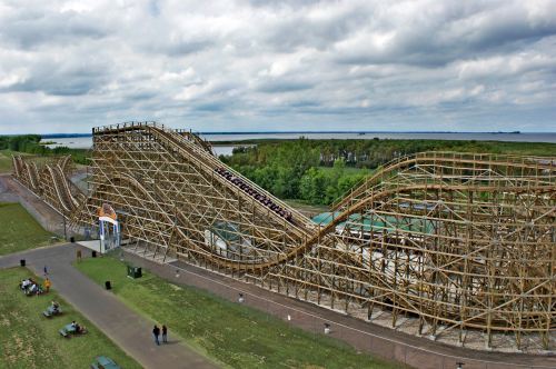 A wooden roller coaster with steep hills and curves, set against a cloudy sky and green landscape.