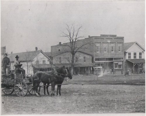 Black and white photo of two men on a horse-drawn cart in a rural town, with buildings in the background.