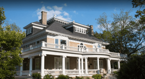 A grand, historic mansion with a large porch, white columns, and lush greenery under a blue sky.