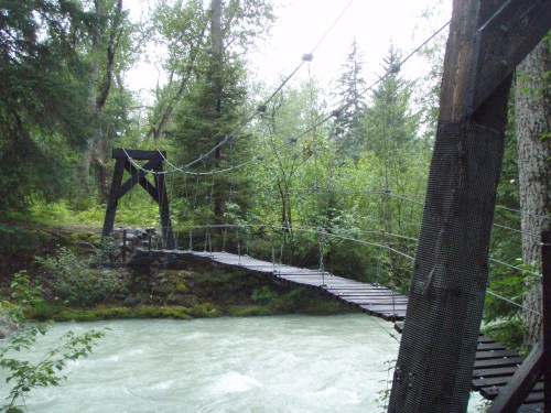A wooden suspension bridge spans a rushing river, surrounded by lush green trees and a cloudy sky.