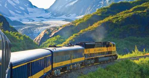 A yellow Alaska train travels through a scenic landscape with mountains and a glacier in the background.