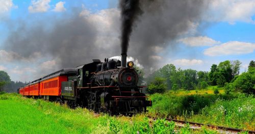 A vintage steam locomotive chugs along a green landscape, billowing black smoke against a blue sky with fluffy clouds.