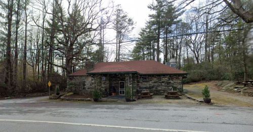 A stone building with a red roof, surrounded by trees, located by a road with picnic tables outside.