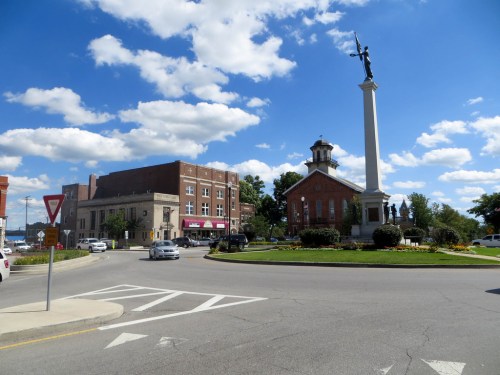 A roundabout featuring a monument, surrounded by buildings and a blue sky with fluffy clouds.