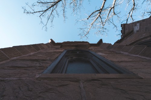 View looking up at a stone building with a window, framed by bare branches against a clear blue sky.