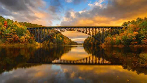 A steel bridge arches over a calm river, surrounded by vibrant autumn foliage and a colorful sunset sky.