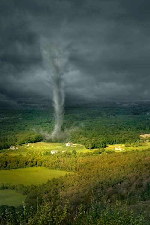 A dramatic landscape featuring a tornado forming over green fields and distant houses under a dark, cloudy sky.
