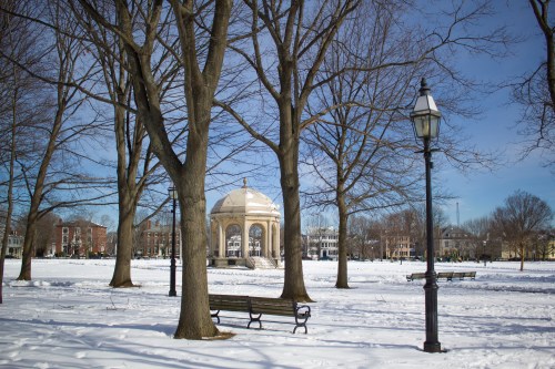 A snow-covered park with bare trees, a gazebo in the background, and a bench beside a lamp post under a clear blue sky.