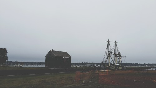 A cloudy day at a harbor featuring a historic building and a tall ship docked nearby.