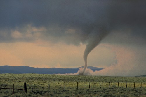 A tornado forms over a grassy field, with dark clouds and distant mountains in the background.
