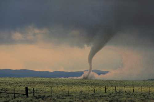 A tornado forms over a grassy field, with dark clouds and distant mountains in the background.