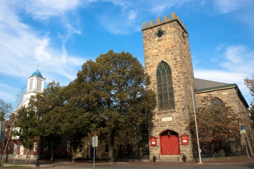Historic stone church with a tall tower, surrounded by trees and a blue sky with scattered clouds.