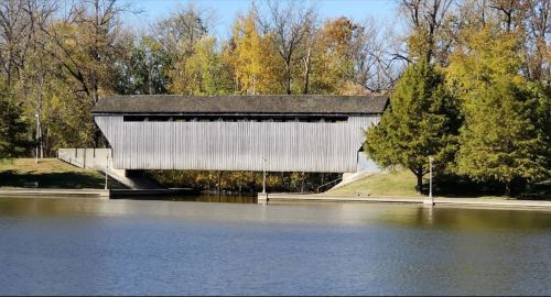 A wooden covered bridge spans a calm lake, surrounded by trees with autumn foliage.