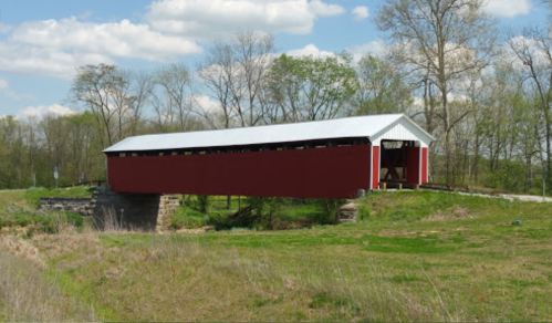 A red covered bridge spans a grassy area, surrounded by trees and a blue sky with fluffy clouds.