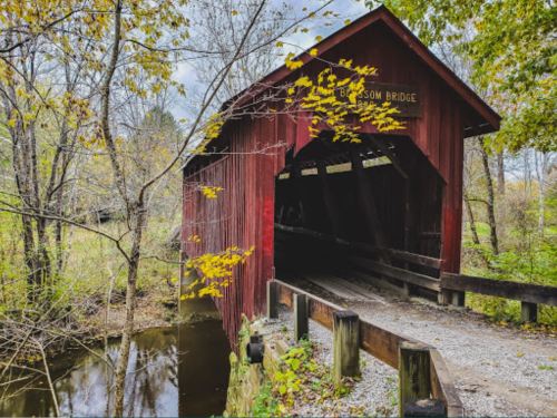 A red covered bridge spans a small stream, surrounded by trees with autumn leaves. Overcast sky in the background.