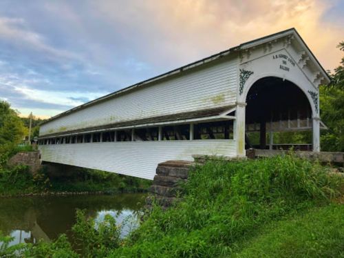 A historic covered bridge spans a calm river, surrounded by lush greenery and a colorful sky at sunset.
