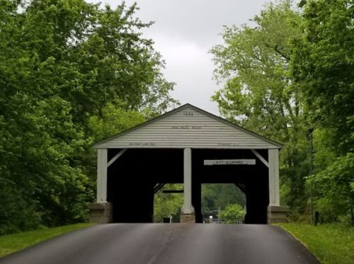 A covered bridge surrounded by lush green trees, leading to a road under a cloudy sky.