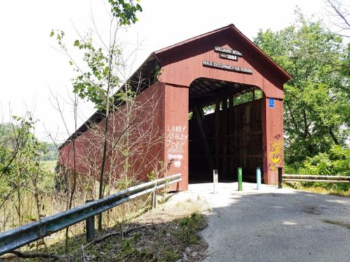 A red covered bridge surrounded by greenery, with graffiti on the side and a gravel path leading to it.