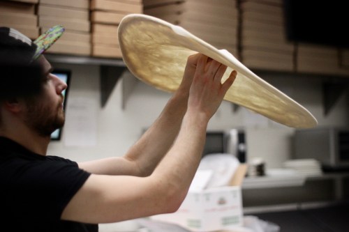 A person in a black shirt stretches pizza dough high above their head in a kitchen setting.