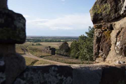 View through stone ruins overlooking a vast landscape with fields and distant trees under a clear blue sky.