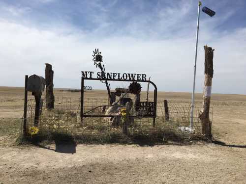 Sign for Mt. Sunflower, the highest point in Kansas, with a metal gate and a flagpole against a vast, open landscape.