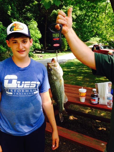 A young boy stands next to a person holding a large fish on a scale, with trees and a picnic area in the background.