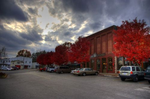 A quaint town street lined with vibrant red trees under a cloudy sky, featuring brick buildings and parked cars.