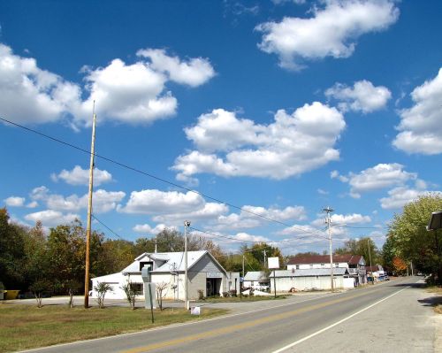 A rural road lined with small buildings under a bright blue sky filled with fluffy white clouds.