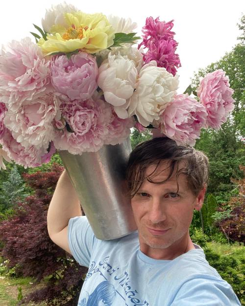 A person holds a large bucket overflowing with colorful peonies in a garden setting.