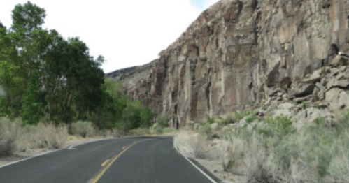 A winding road alongside tall rock cliffs and sparse vegetation under a cloudy sky.