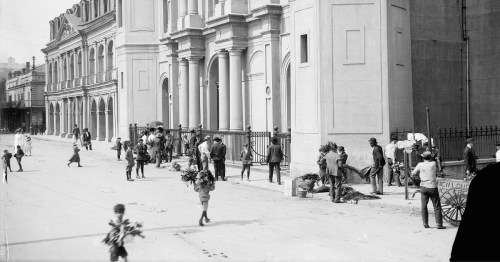 Black and white historical street scene with people selling flowers and engaging in various activities near a grand building.