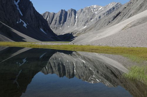 A serene mountain landscape with steep cliffs reflected in a calm lake, surrounded by rocky terrain and patches of grass.
