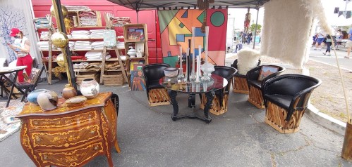 A market stall featuring vintage furniture, including a glass table and ornate chairs, with colorful artwork in the background.