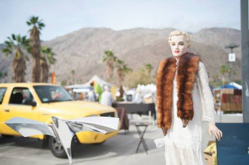 A vintage mannequin in a fur stole stands near a yellow taxi at an outdoor market with mountains in the background.