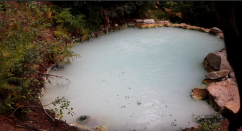 A serene, light blue natural hot spring surrounded by rocks and lush greenery.