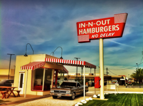 A vintage In-N-Out Burgers location with a classic car parked outside and a bright red sign against a blue sky.